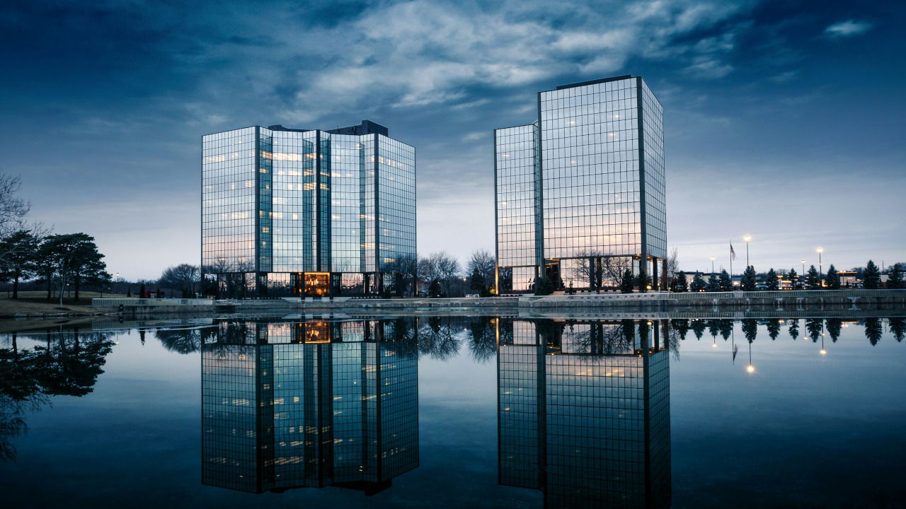 Two mirrored office buildings reflected in a still pool at dusk, with a moody sky overhead. The calm surface and looming glass towers evoke quiet tension and the polished yet isolating nature of corporate life.