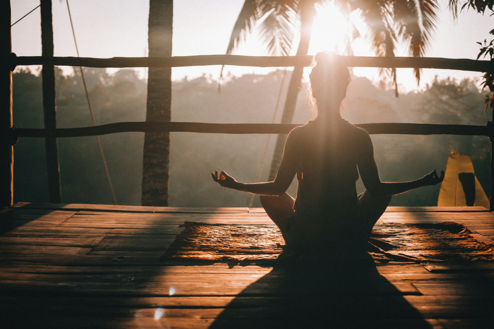 A woman sits cross-legged in a meditative pose on a wooden deck at sunrise, silhouetted by warm sunlight filtering through palm trees. Her hands rest on her knees in a gesture of calm and mindfulness.