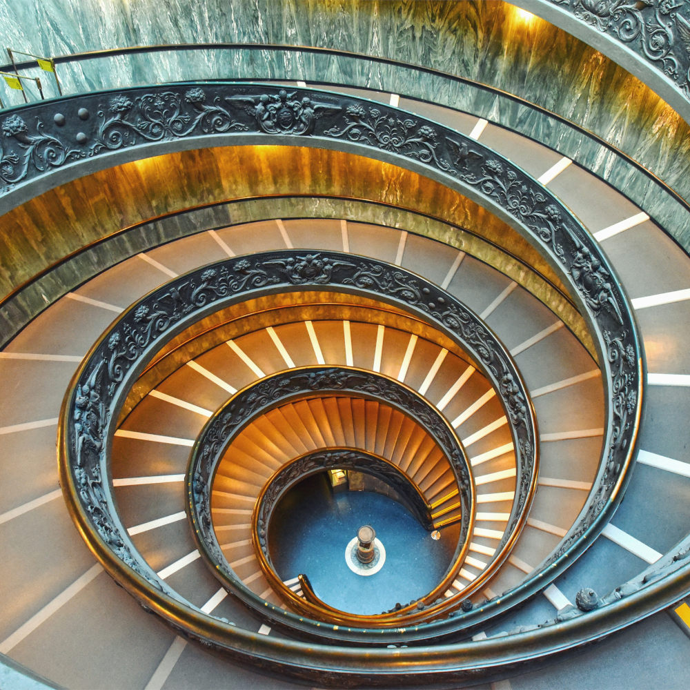 A top-down view of a grand spiral staircase forming a Fibonacci-like spiral, featuring ornate dark railings, golden lighting, and marble walls, leading to a circular landing below.