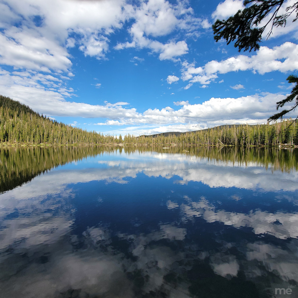 A calm alpine lake in Colorado reflects a vivid blue sky with scattered white clouds and surrounding pine-covered mountains, creating a nearly perfect mirror image.
