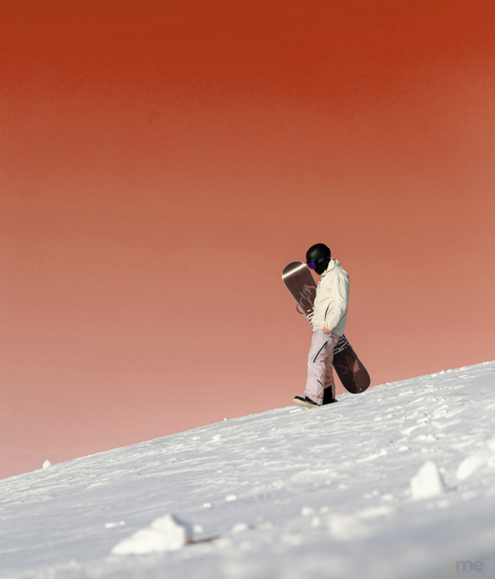 A lone snowboarder in a white jacket and helmet walks down a snowy slope, carrying their snowboard at their side, against a dramatic red-orange sky.