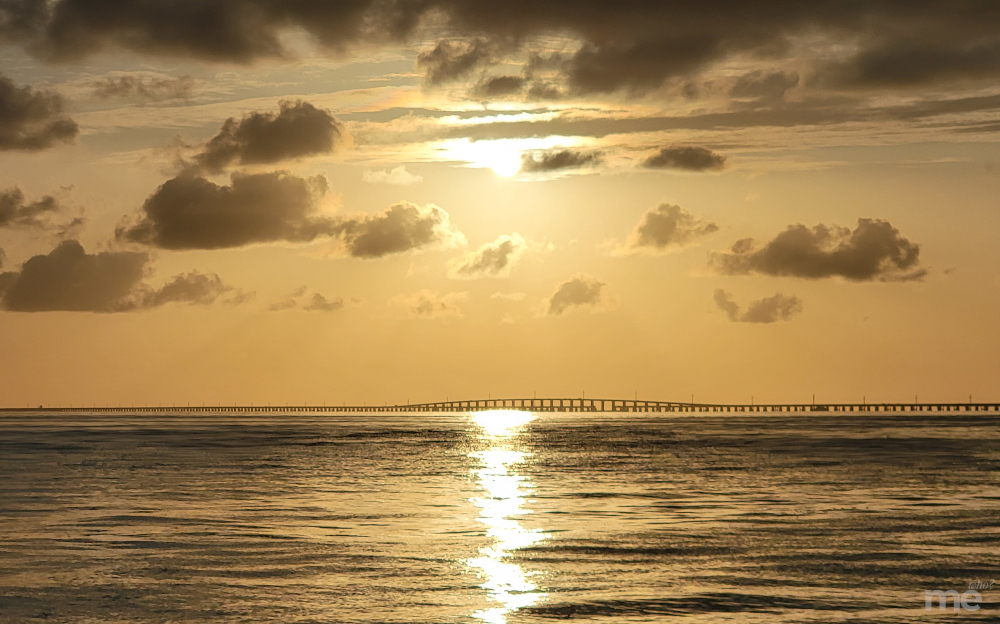A golden sunset with the sun peeking through scattered clouds, reflecting across calm ocean water with a distant bridge on the horizon.