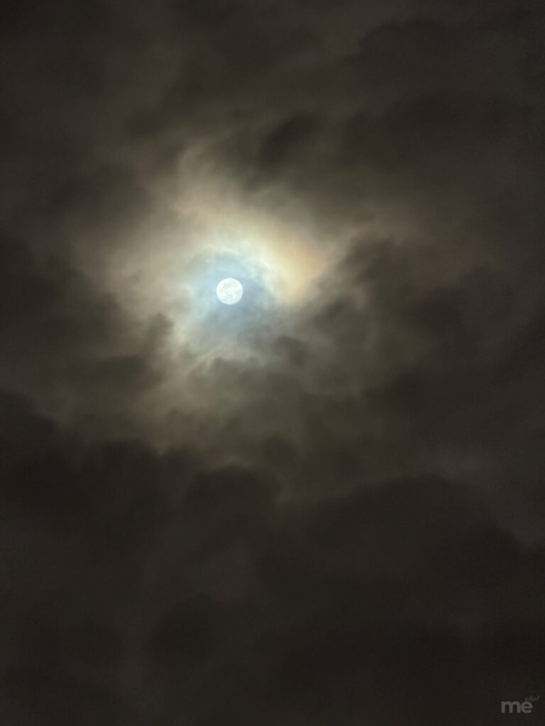 A bright moon glows through thick, dark clouds, surrounded by a faint halo of light in the night sky.