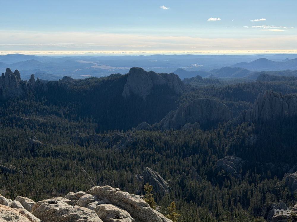 A wide landscape photograph showing forested mountains and rocky outcrops stretching into the distance under a clear blue sky.
