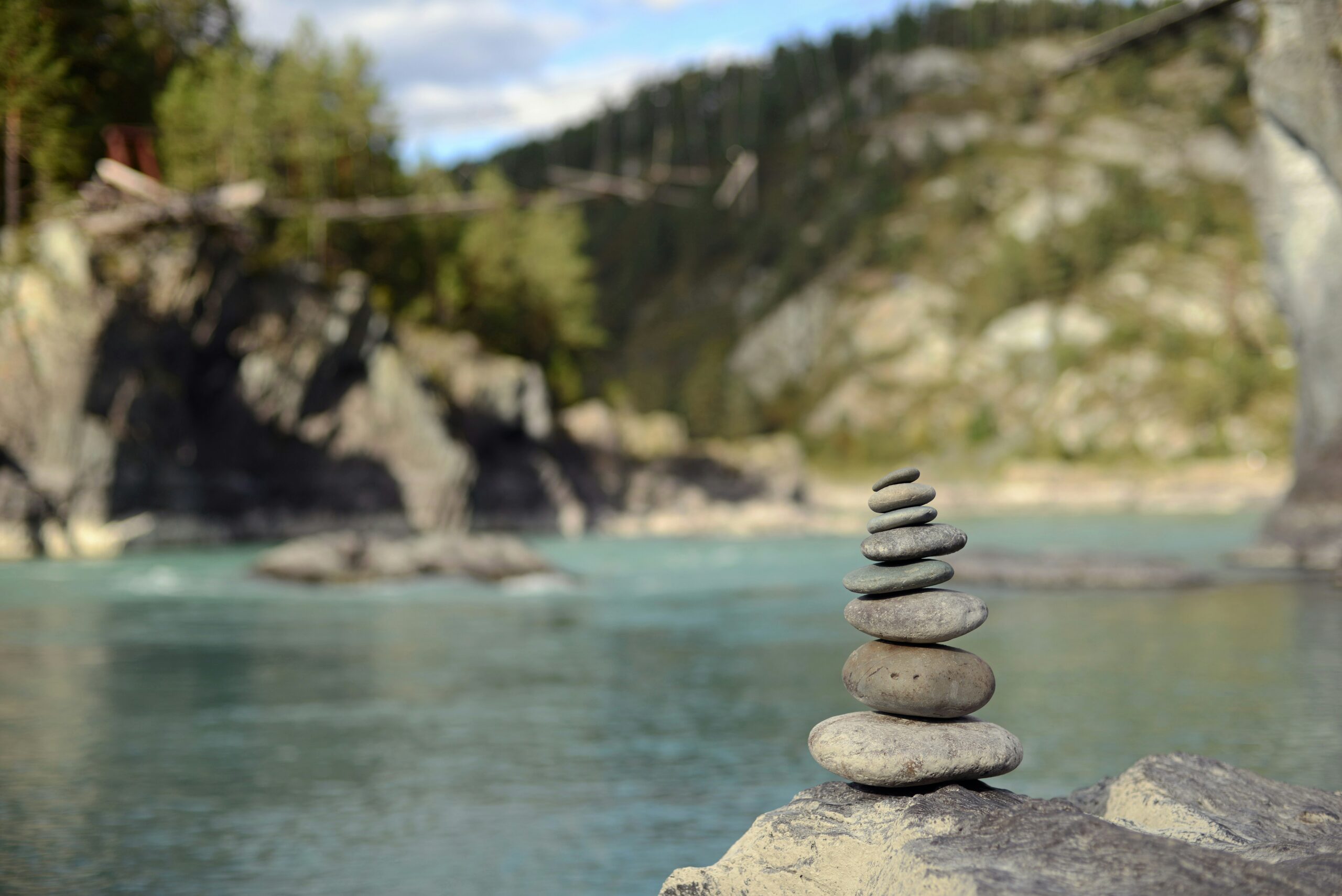 A small stack of smooth stones balanced on a rock beside a calm river, with a forested canyon blurred in the background.