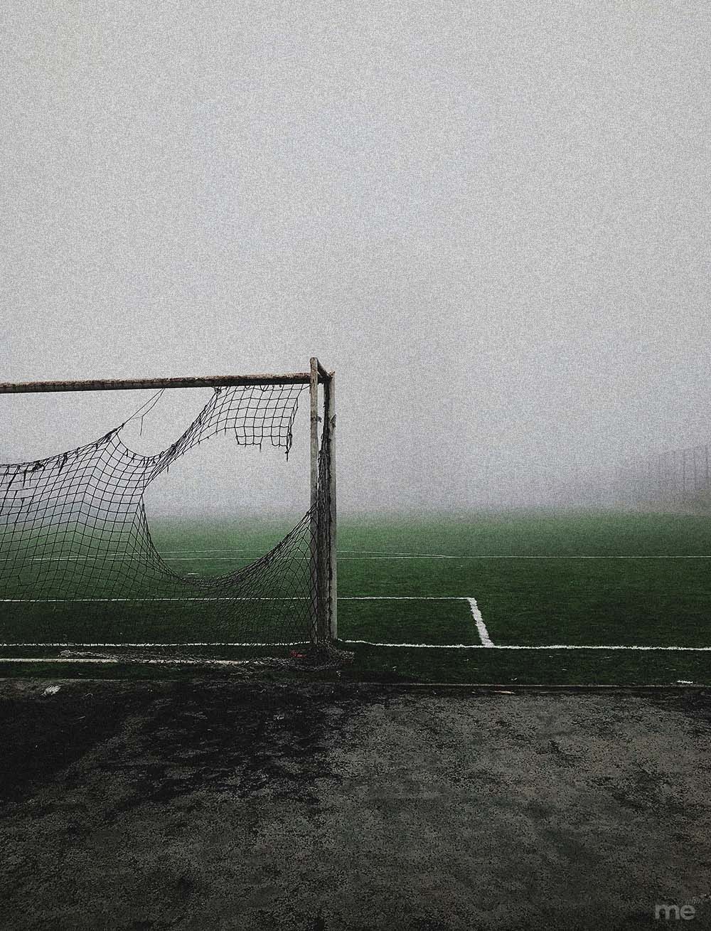 A vertical, moody photograph of a weathered soccer goal with a severely torn net standing on a green field. Thick, grey fog fills the background, completely obscuring the horizon and creating an atmospheric, desolate scene.