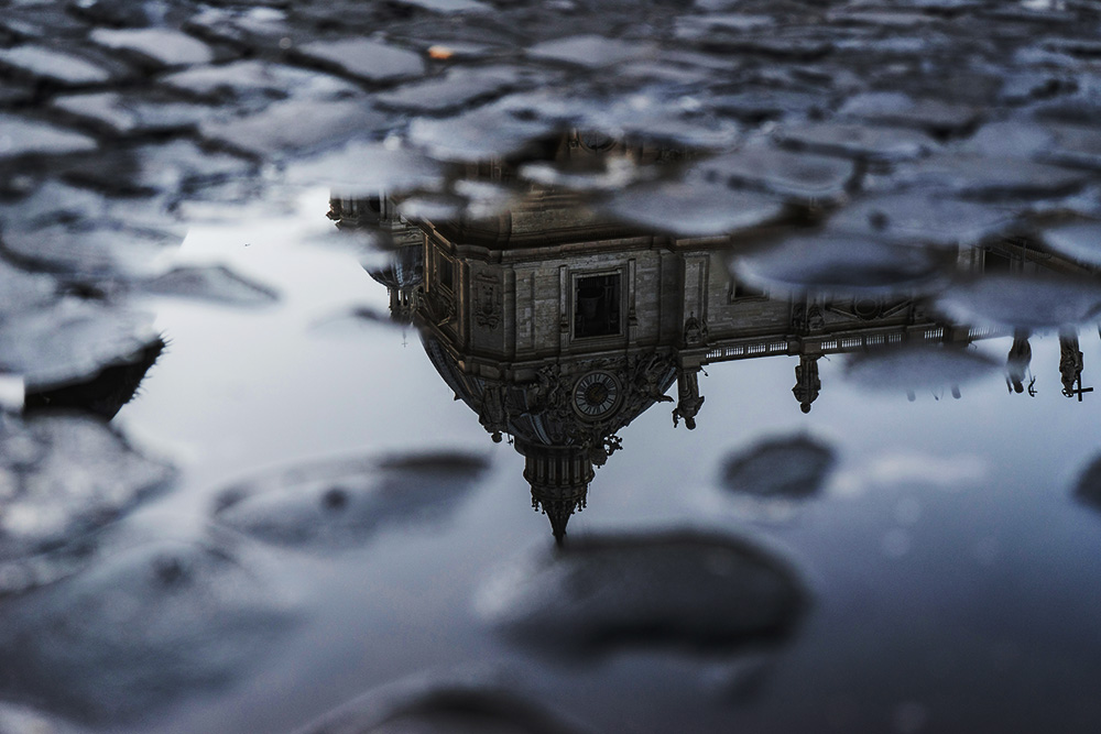 A moody, low-angle photograph of a cobblestone street where a large puddle perfectly reflects a grand, classical building. The reflection shows an ornate dome, a clock tower, and several statues against a gray sky. The surrounding wet cobblestones are out of focus, creating a soft bokeh effect in the foreground and background.