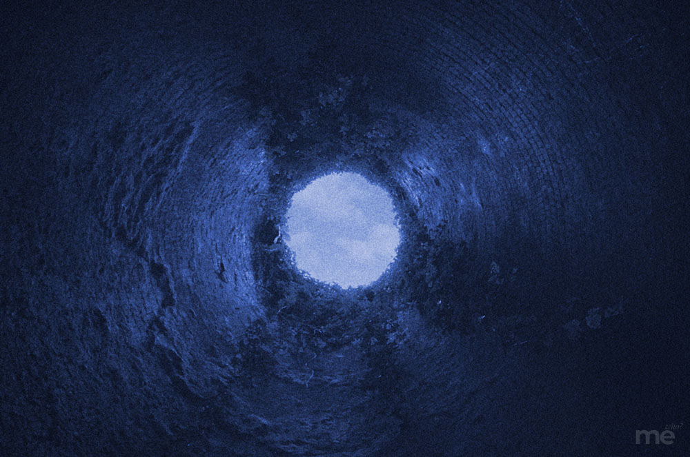 A grainy, deep-blue toned photograph looking straight up from the bottom of a dark, circular tunnel toward a bright opening of light at the top.