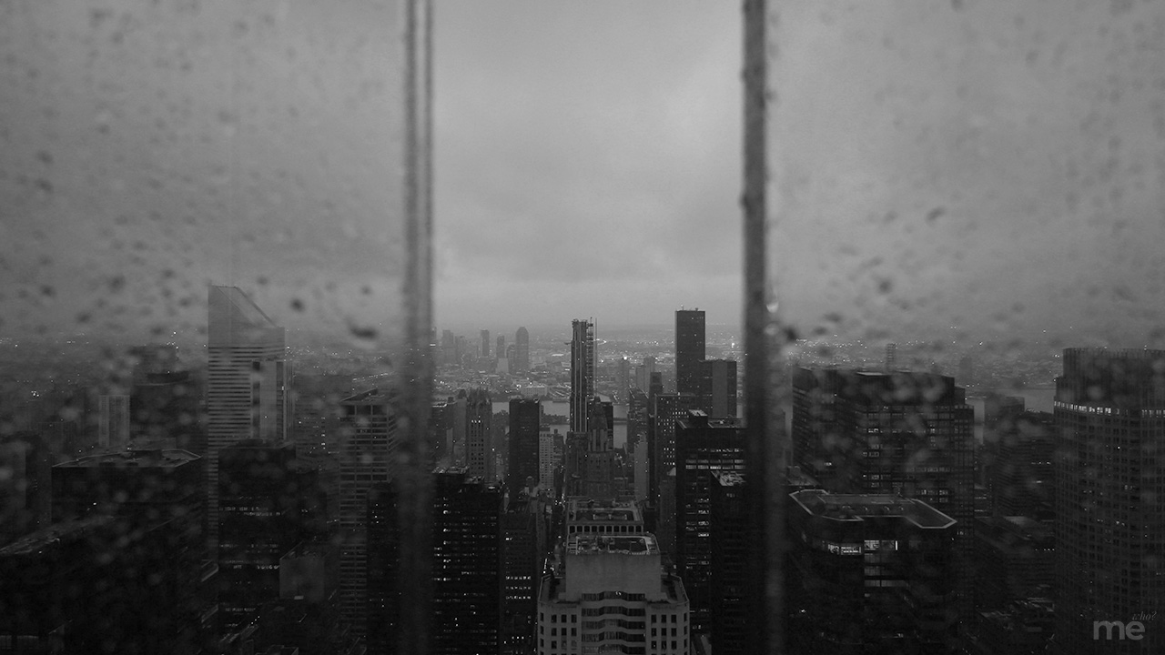 A moody, black-and-white cityscape viewed through a window covered in raindrops. The tall skyscrapers of a dense urban skyline are partially obscured by the condensation and heavy, overcast clouds. Two vertical streaks of water on the glass frame the center of the city, creating a sense of depth and isolation.