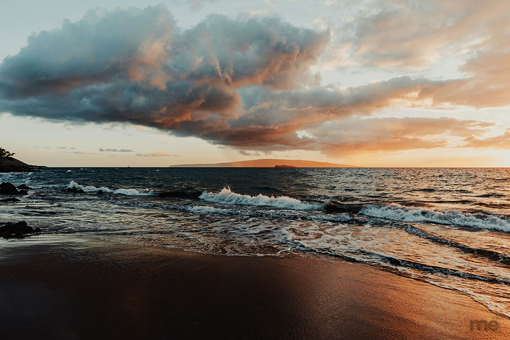 Waves washing onto a dark sand beach at sunset with large, colorful storm clouds overhead and a distant island in the background.