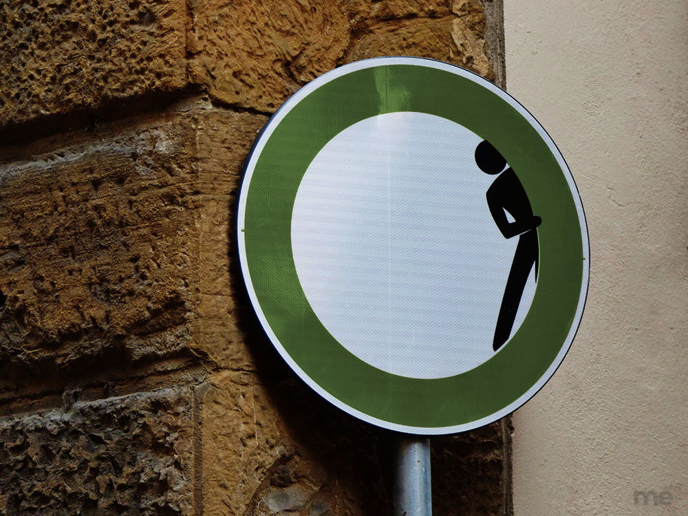 A playful piece of street art featuring a circular sign with a thick green border and a white center. A black silhouette of a person is positioned to look as if they are peeking out from behind the green ring into the white void. The sign is mounted on a metal pole against a rustic, textured brown stone wall.