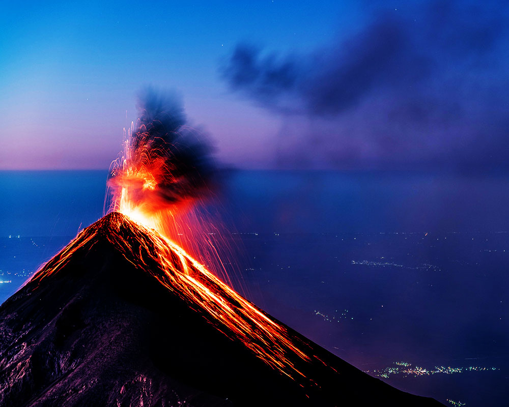 A dramatic night photograph of a volcanic eruption. Molten red lava flows down the steep, dark slopes of the peak while a vibrant explosion of fire and sparks bursts from the crater. A plume of dark smoke rises into a deep blue and purple twilight sky.