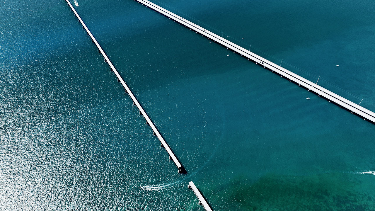 A high-angle aerial view of two long, white bridges stretching across a vast expanse of deep blue water. One bridge is continuous, while the other appears to have a gap where a small boat is passing through.