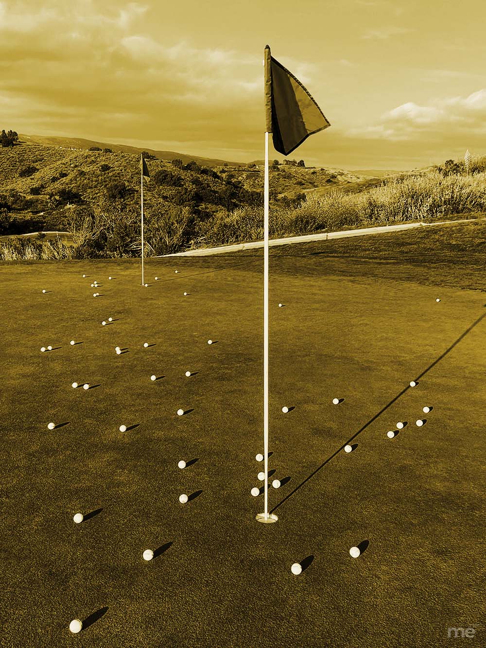 A sepia-toned high-angle shot of a golf putting green littered with golf balls surrounding a flagstick, with rolling hills in the background.