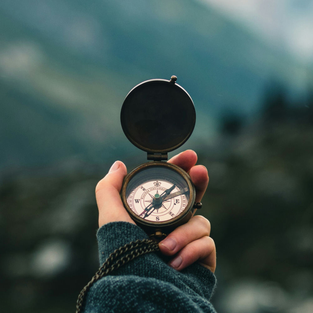 Close-up of a person’s hand holding an antique brass compass against a blurred mountain backdrop, symbolizing inner guidance and clarity of direction.