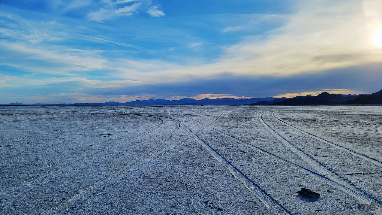 A wide landscape shot of a vast salt flat at dusk. Several crisscrossing tire tracks mark the textured, white ground, leading toward a distant range of blue mountains under a sprawling sky with soft blue and yellow hues.