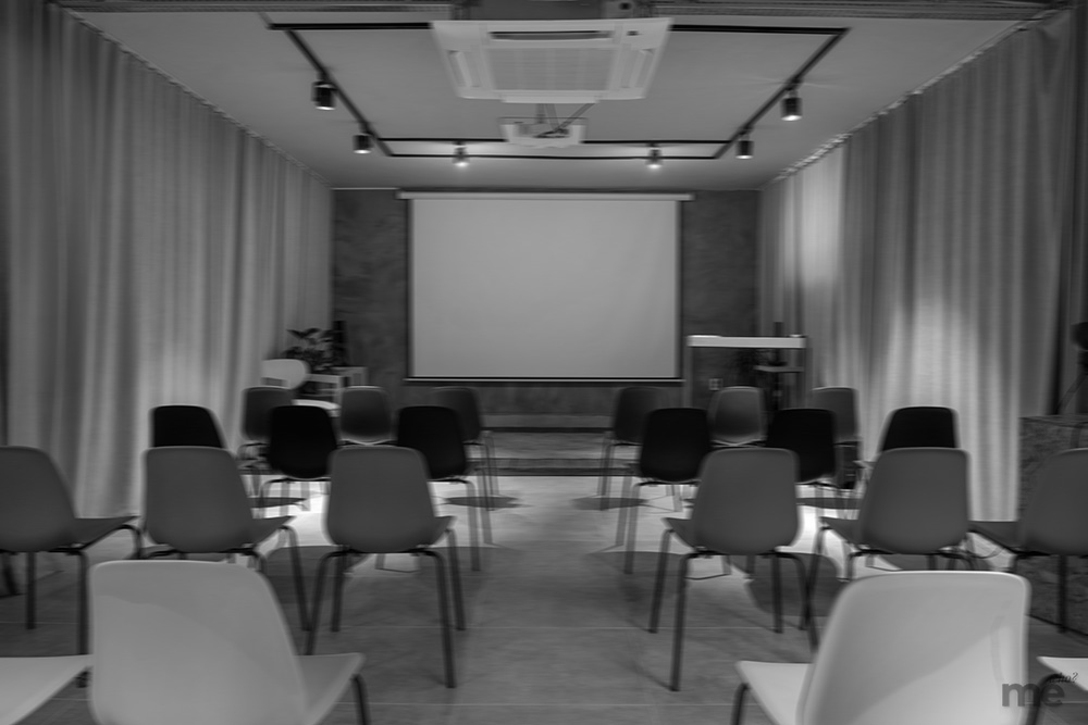 A black and white photograph of an empty, modern presentation room with rows of chairs facing a large blank projector screen and track lighting.