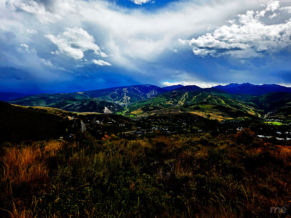 A high-vibrancy landscape photograph overlooking a lush green valley and rolling mountain ranges under a dramatic sky. Massive white and dark blue storm clouds tower over the peaks, while the foreground shows sunlit golden grasses and shrubs.