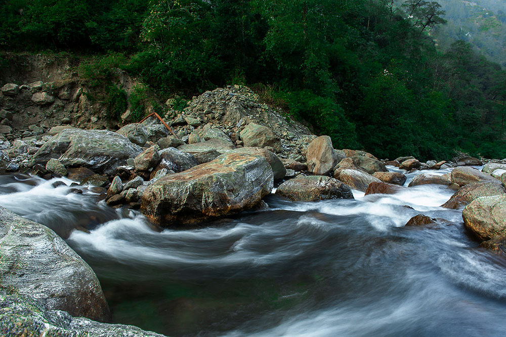 A long-exposure photograph of a mountain river rushing over large, mossy grey boulders. The moving water appears as a smooth, silky white mist due to the slow shutter speed. In the background, a steep hillside is covered in dense, dark green foliage and trees.