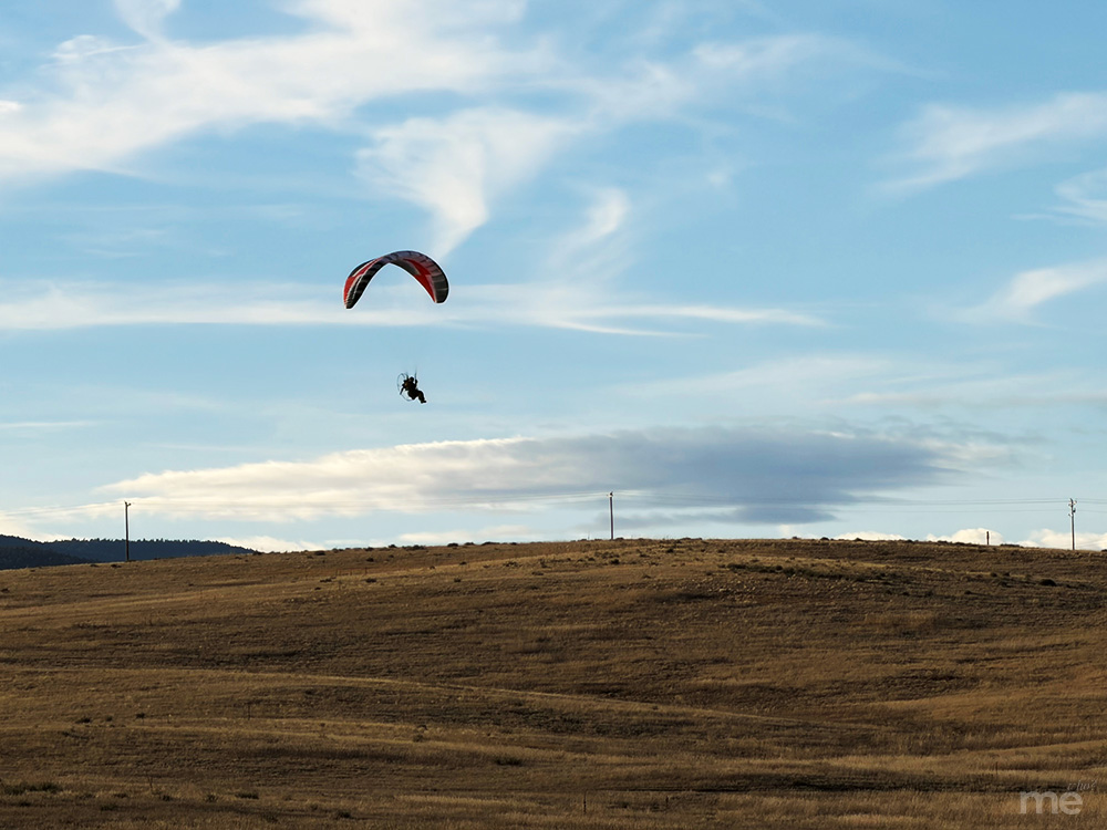 A wide-angle photograph of a powered paraglider soaring through a bright, hazy blue sky with wispy white clouds. The paraglider, featuring a red, black, and white canopy, hangs above a vast, rolling golden-brown field.