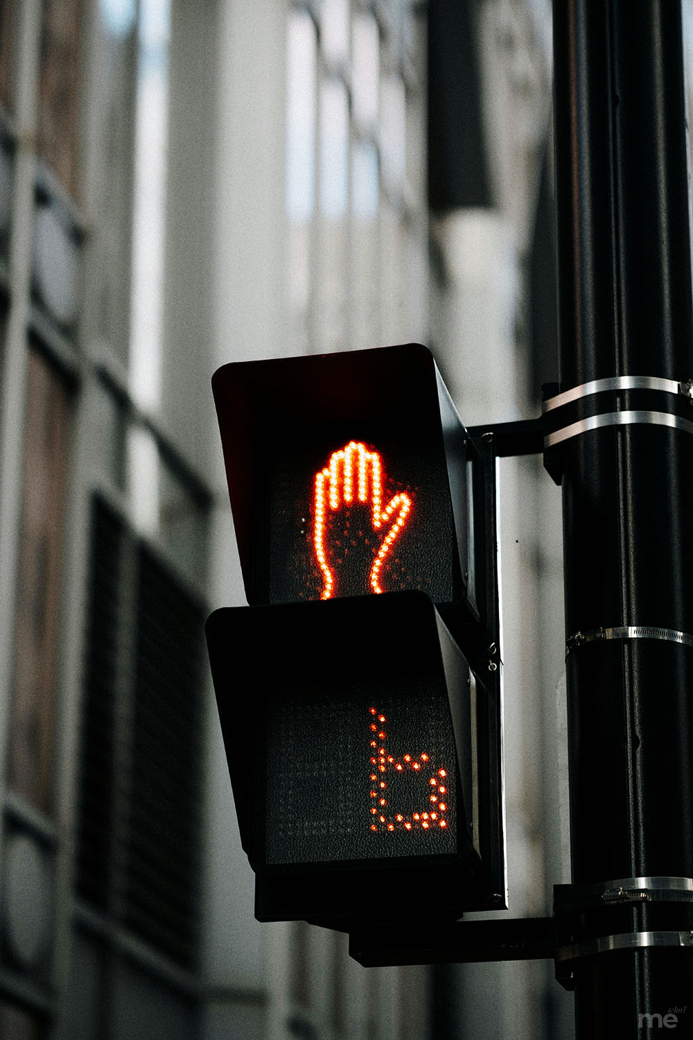 A vertical photograph of a pedestrian crossing signal mounted on a black metal pole. The top portion of the signal displays a glowing orange "don't walk" hand icon, while the bottom section shows a digital countdown timer displaying the number "6." The background features the blurred architecture of city buildings in neutral tones.