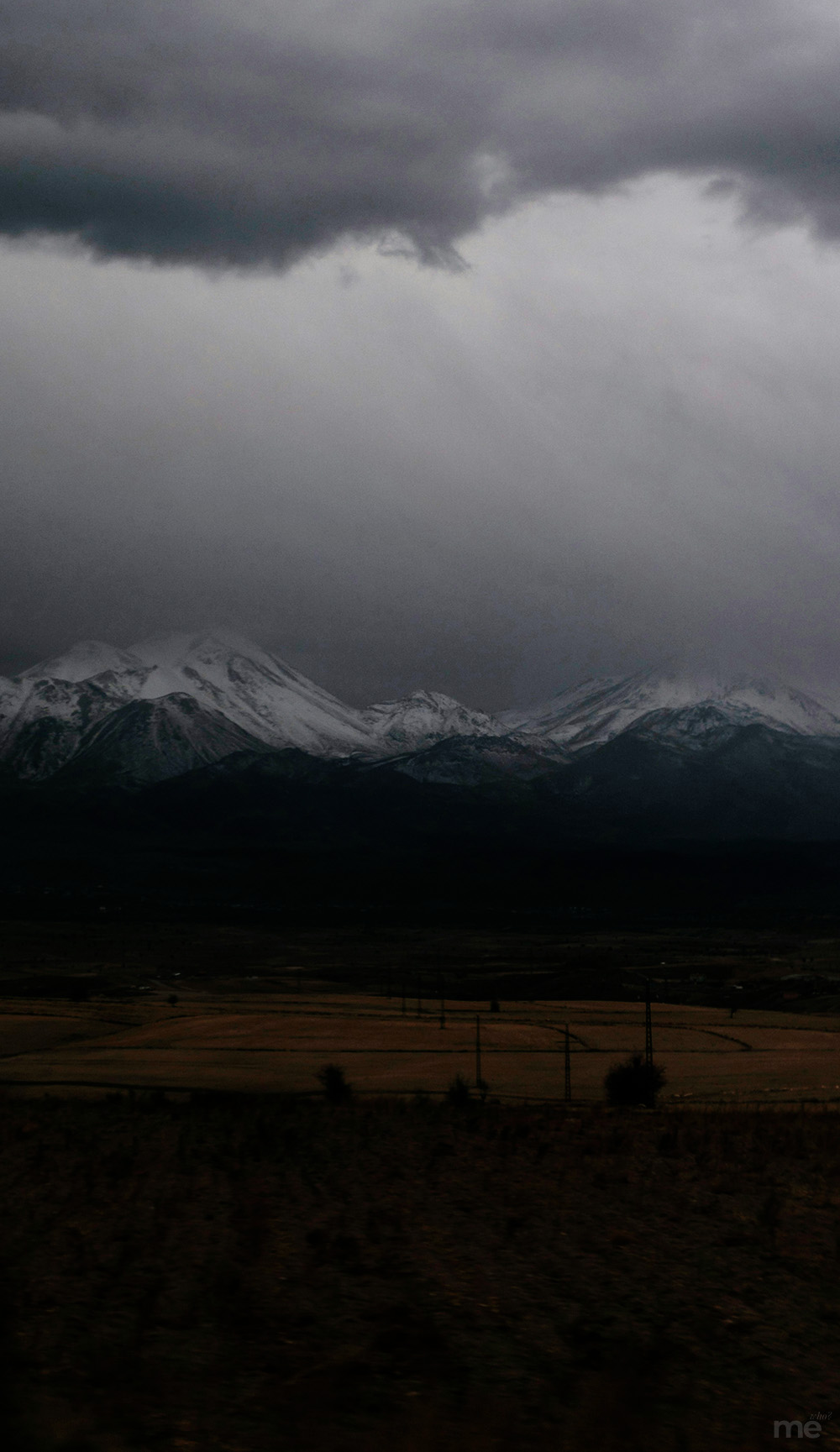 A vertical, low-light landscape photograph of snow-capped mountains under a heavy, dark storm cloud. The jagged peaks are dusted with white snow, contrasting against the deep shadows of the lower slopes.