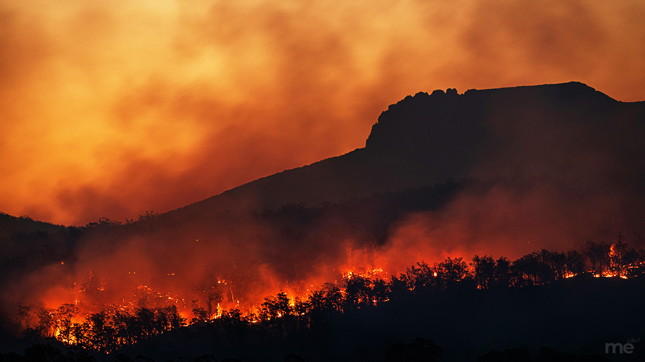 A dramatic landscape photograph of a forest fire raging across a mountainside at night. Glowing red and orange flames consume the treeline in the foreground, sending thick plumes of smoke into an ember-colored sky. A large, dark silhouette of a rugged mountain peak looms in the background against the hazy, fiery atmosphere.