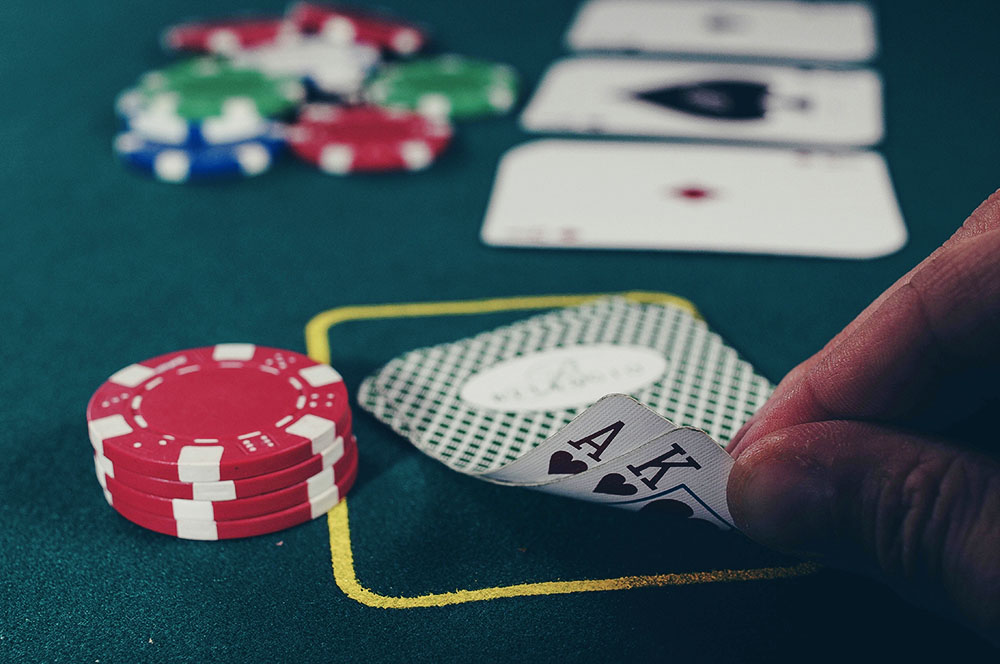 A close-up of a poker player’s hand subtly lifting the corners of two cards to reveal an Ace and a King. A stack of red chips sits nearby on the green felt table, with blurred community cards and additional chips in the background.