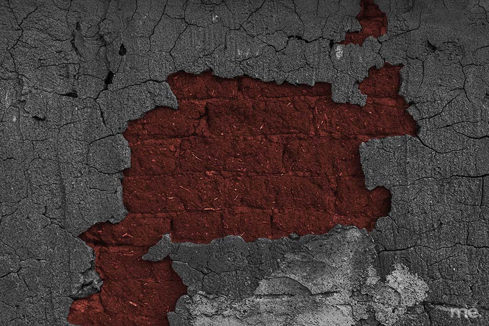 A high-contrast, textured photograph of a crumbling wall. The outer layer consists of dark grey, heavily cracked plaster that has peeled away in a large, irregular section to reveal an underlying layer of deep red brick or clay. The exposed red core has a rough, fibrous texture.