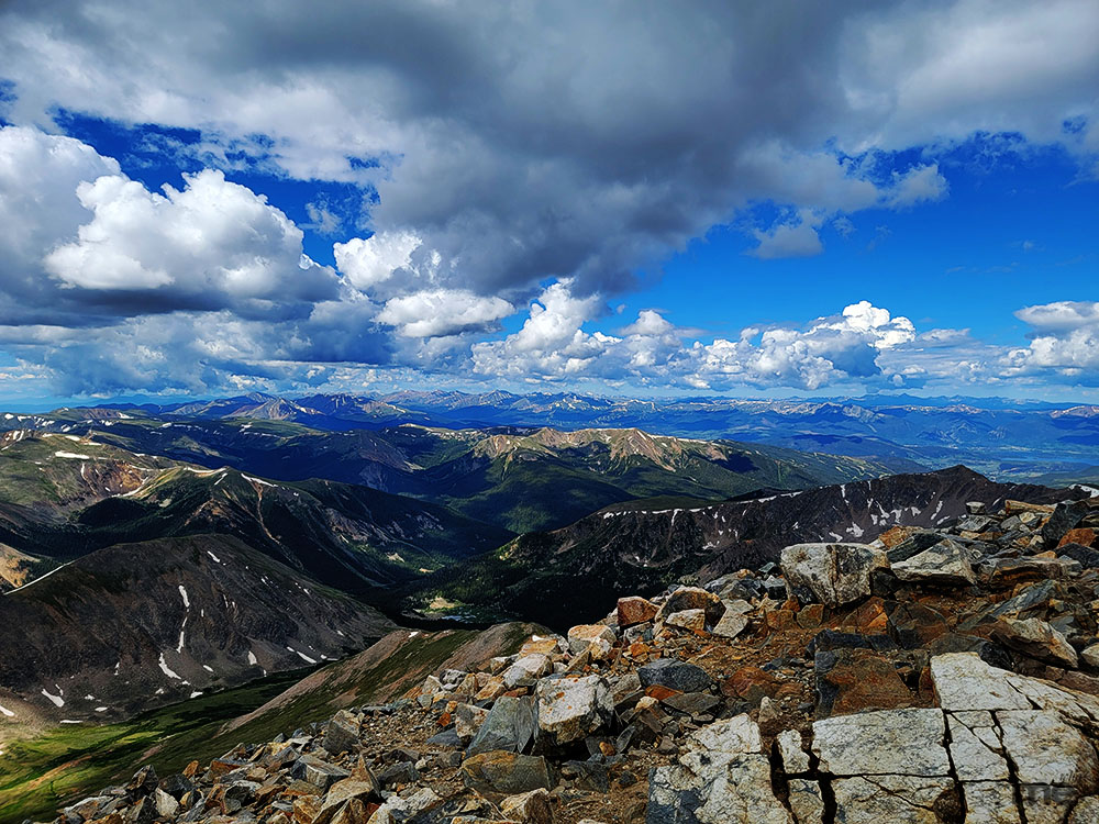 A high-altitude, wide-angle landscape photograph taken from a rocky mountain summit. The foreground is filled with jagged, light-colored boulders and loose scree. In the distance, a vast range of deep blue and green mountain peaks stretches toward the horizon under a bright blue sky filled with dramatic, white cumulus clouds.