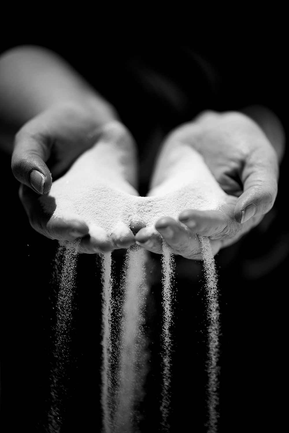 A high-contrast, black-and-white photograph showing a pair of cupped hands holding a mound of fine, white sand. The sand is slipping through the gaps between the fingers, creating several vertical, flowing streams that fall toward the bottom of the frame against a solid black background.