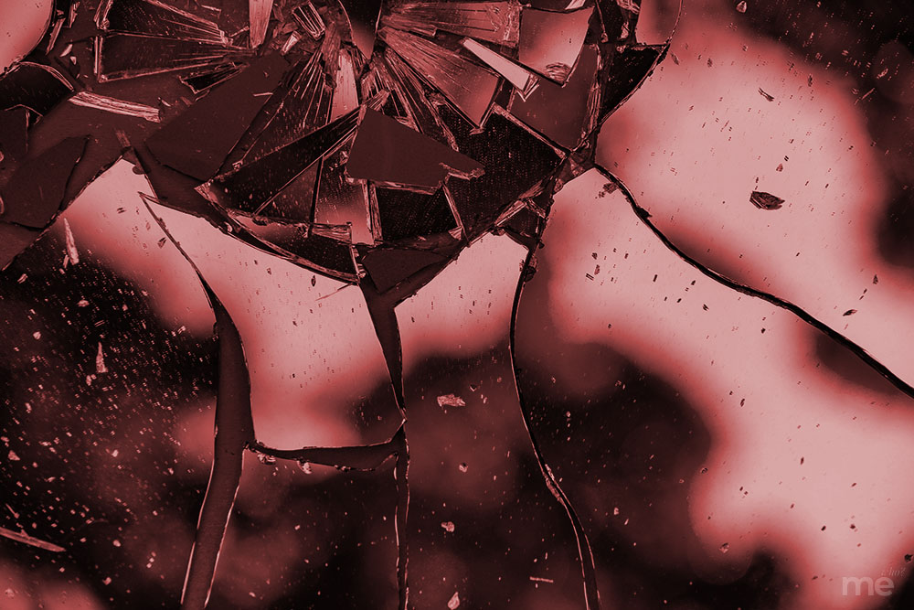 A dramatic, close-up photograph of shattered glass with a deep red and pink color grade. Large, jagged cracks radiate from a central impact point near the top of the frame, where several small shards are missing. The background is softly blurred with dark, indistinct shapes, creating a high-contrast and moody aesthetic.