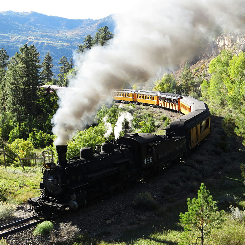 A high-angle photograph of a vintage black steam locomotive pulling a long line of yellow and black passenger cars along a curved track. A thick plume of white and grey smoke billows from the engine's smokestack, trailing over the train.