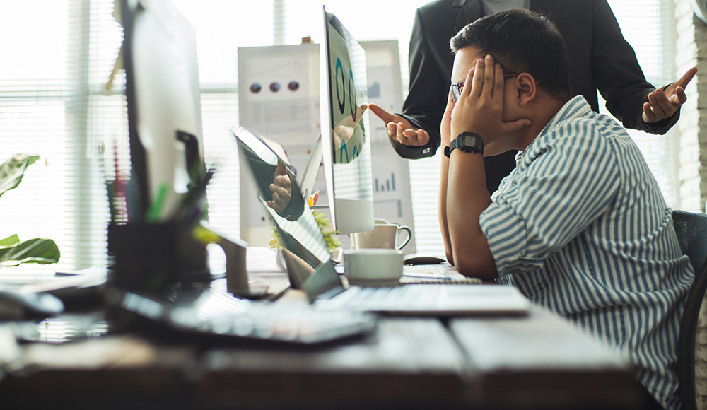 An employee experiencing workplace stress and burnout while receiving feedback from a manager in a modern office setting. The worker appears frustrated, covering his face while sitting in front of a computer monitor.