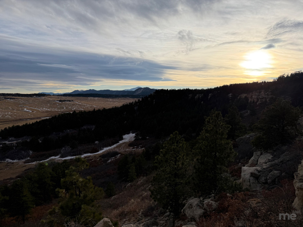 The sun sets over rolling foothills and open plains, with dark pine-covered ridges in the foreground and distant mountains beneath a cloud-streaked sky.