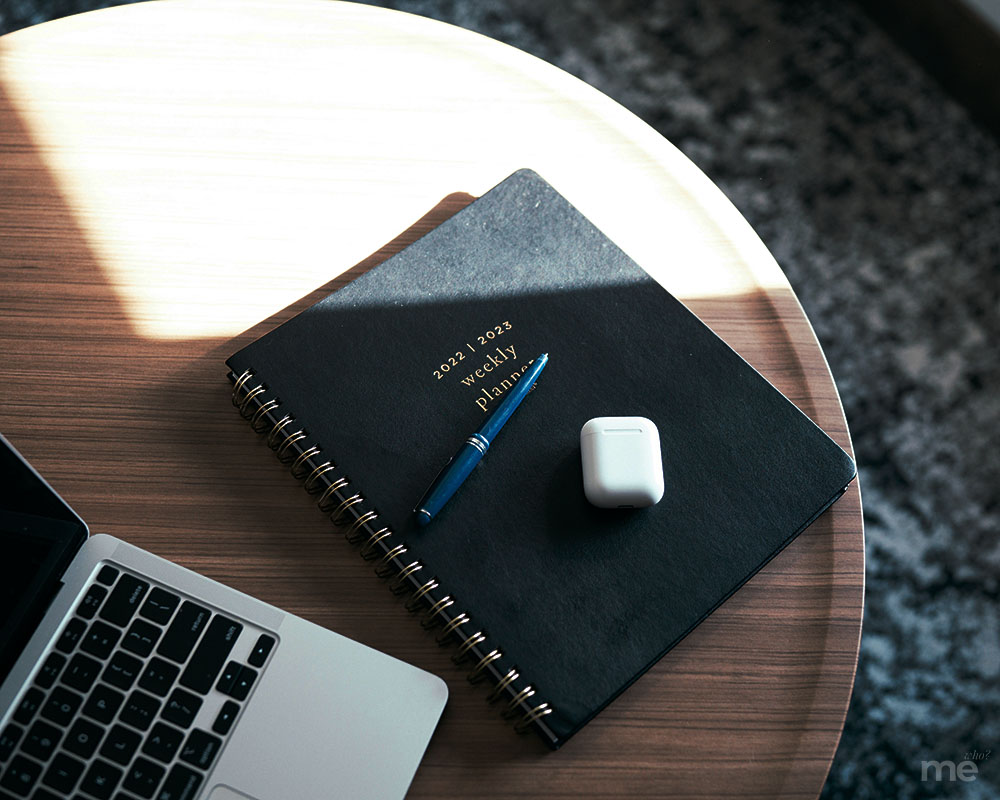 An overhead, angled shot of a workspace on a round wooden table. A black spiral-bound planner sits in the center with a blue pen and a white wireless earbud case resting on its cover. To the left, a silver laptop is partially visible, and a bright beam of sunlight cuts across the table and the notebook.